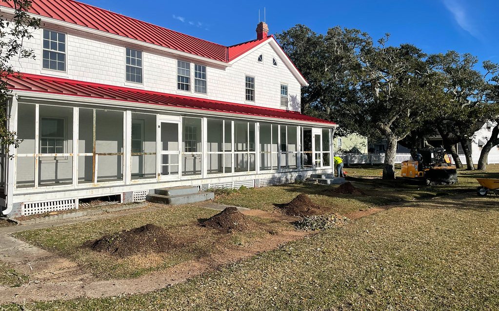 A crew removing a tree stump on the lawn of a large commercial building shaded by mature oaks