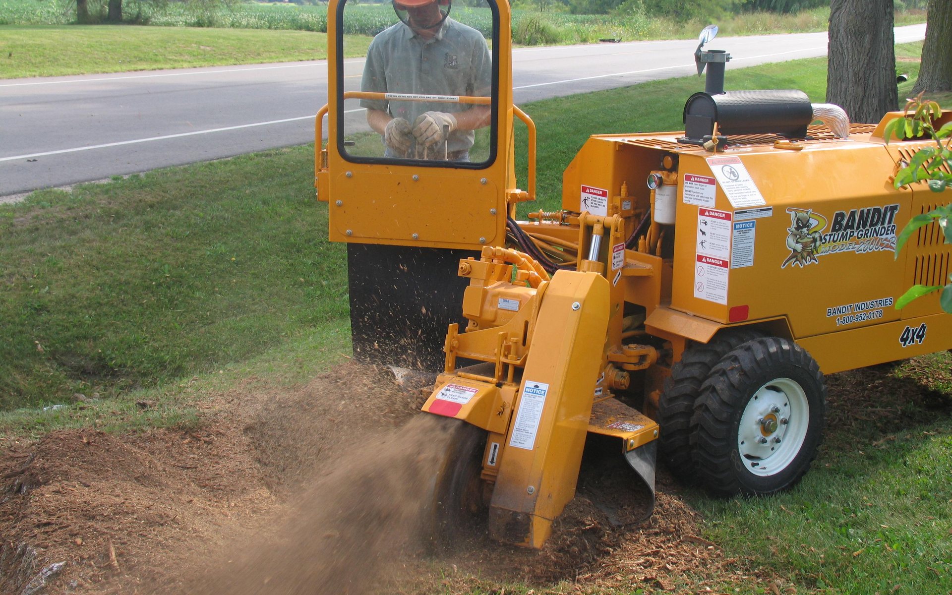 An operator using a stump grinder to grind a stump beside a road, wood chips flying